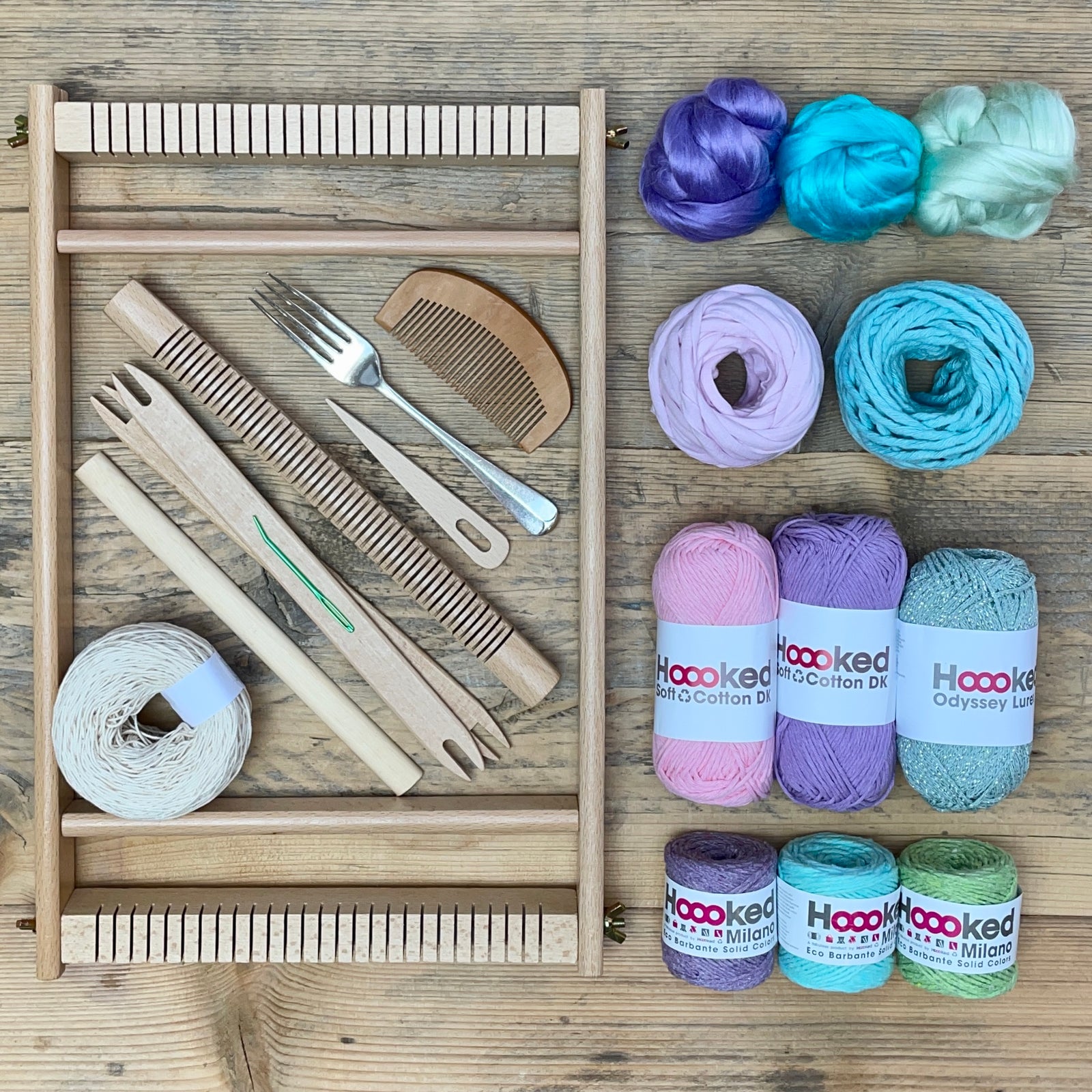 A beginners frame/ tapestry loom weaving starter kit displayed with weaving supplies including assorted yarns in an 'Borealis' colour way and weaving tools.