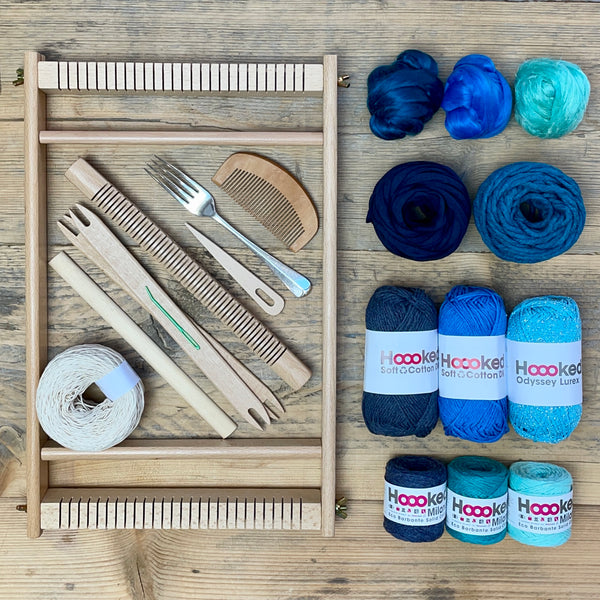 A beginners frame/ tapestry loom weaving starter kit displayed with weaving supplies including assorted yarns in an 'Marine' colour way and weaving tools.