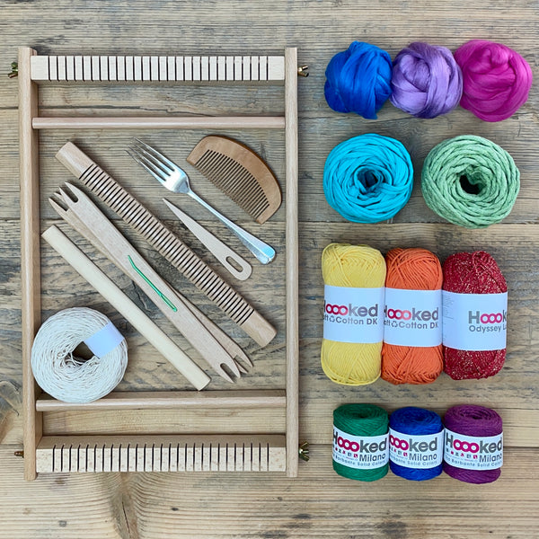 A beginners frame/ tapestry loom weaving starter kit displayed with weaving supplies including assorted yarns in an 'Rainbow' colour way and weaving tools.