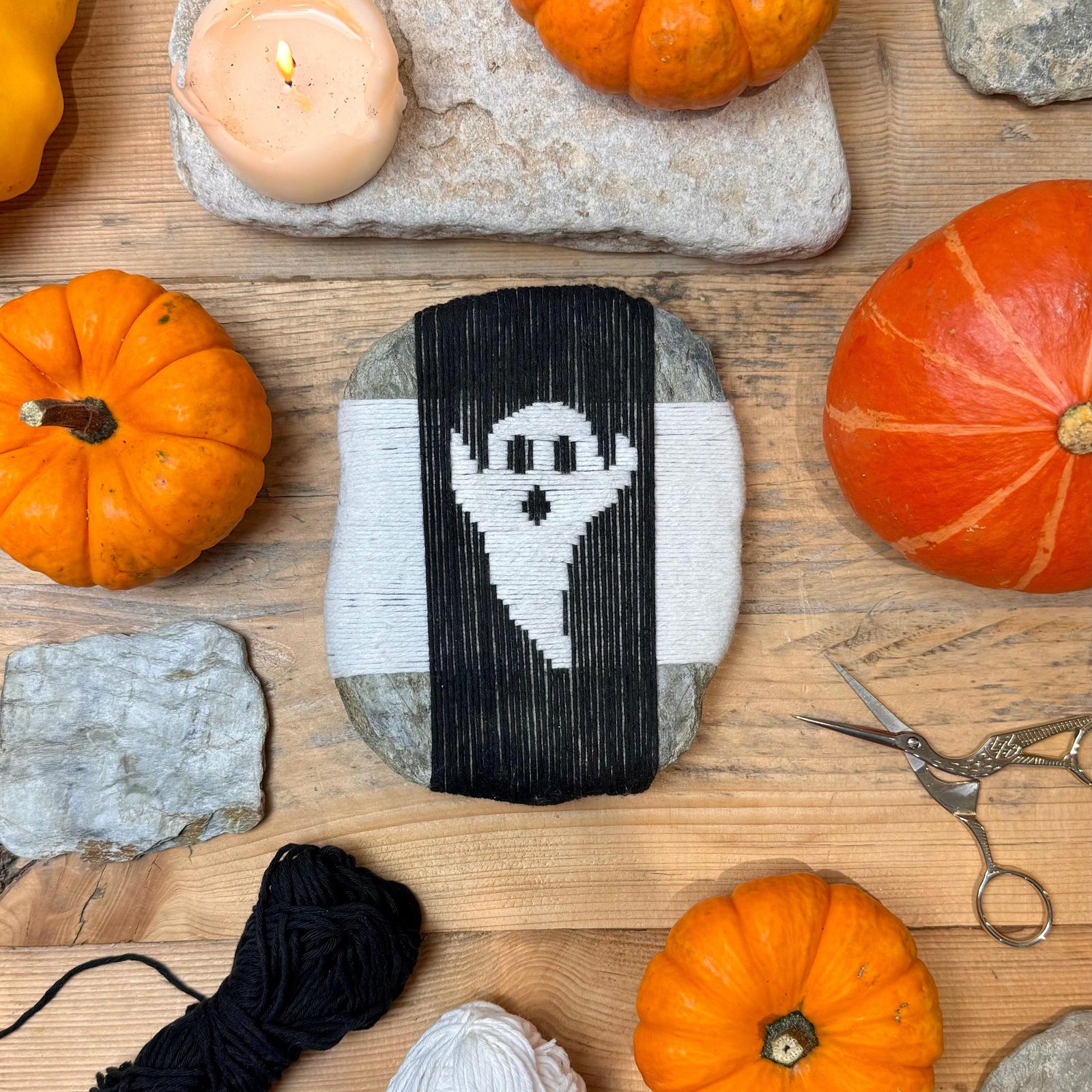 A woven ghost design on a flat stone, surrounded by mini pumpkins, gourds and a lit candle for Halloween styling