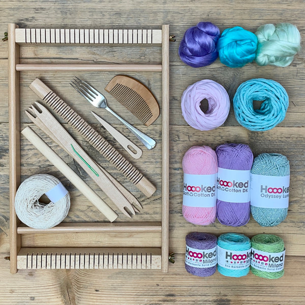 A beginners frame/ tapestry loom weaving starter kit displayed with weaving supplies including assorted yarns in an 'Borealis' colour way and weaving tools.