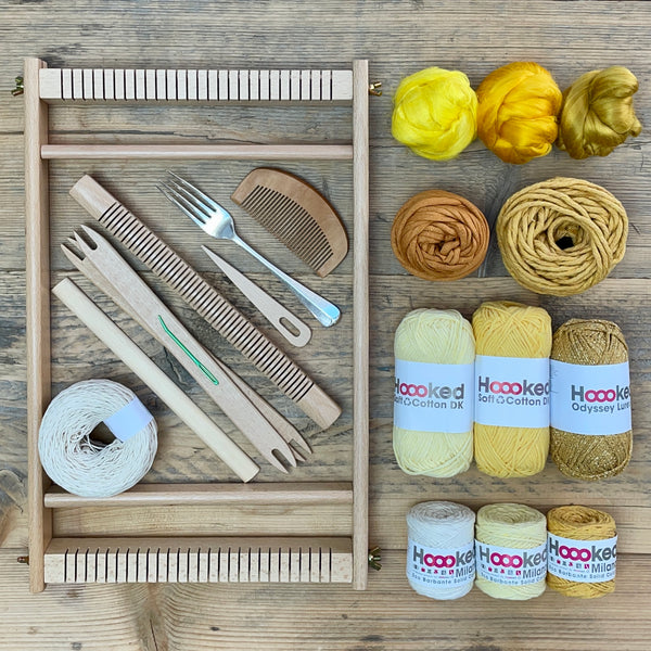 A beginners frame/ tapestry loom weaving starter kit displayed with weaving supplies including assorted yarns in an 'Golden Solstice' colour way and weaving tools.