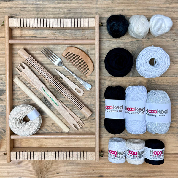 A beginners frame/ tapestry loom weaving starter kit displayed with weaving supplies including assorted yarns in an 'Monochrome Magic' colour way and weaving tools.