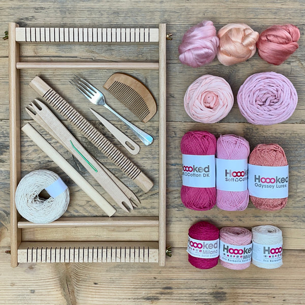 A beginners frame/ tapestry loom weaving starter kit displayed with weaving supplies including assorted yarns in an 'Peach Melba' colour way and weaving tools.