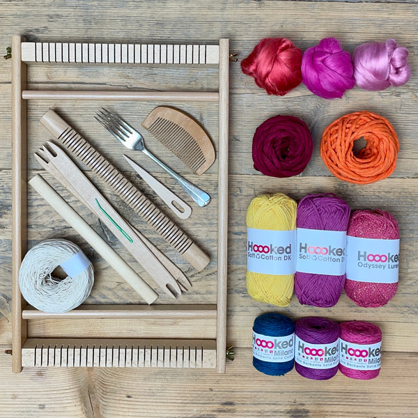 A beginners frame/ tapestry loom weaving starter kit displayed with weaving supplies including assorted yarns in a 'Sunset' colour way and weaving tools.